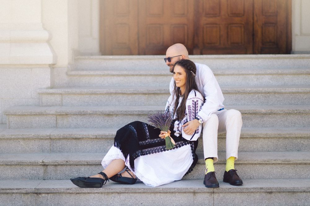 Two people in traditional Romanian folk costumes sitting on stone steps with a casual pose.