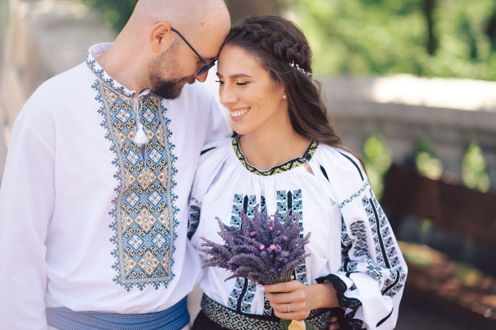 Couple in embroidered Romanian folk costumes holding purple flowers in an outdoor setting.