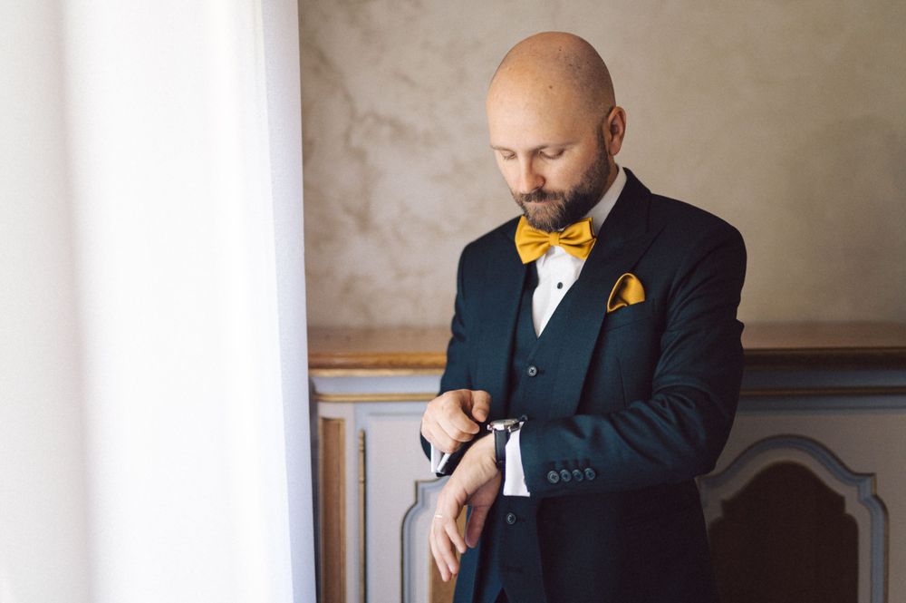 Groom in navy suit with yellow bowtie checks his watch while getting ready.