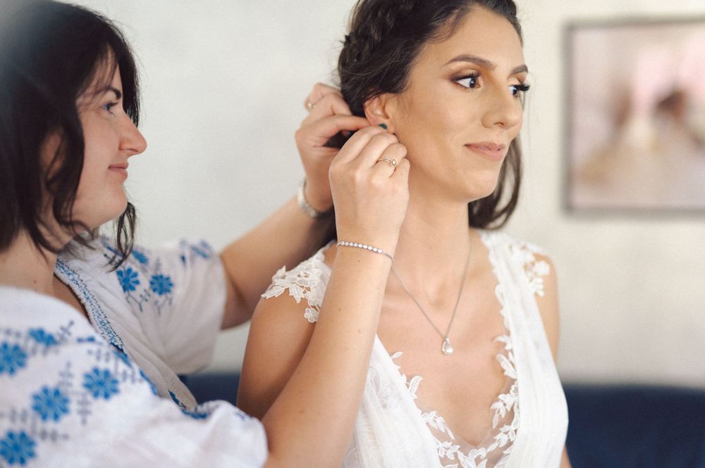 Helper assists with jewelry as bride gets ready in lace wedding gown.
