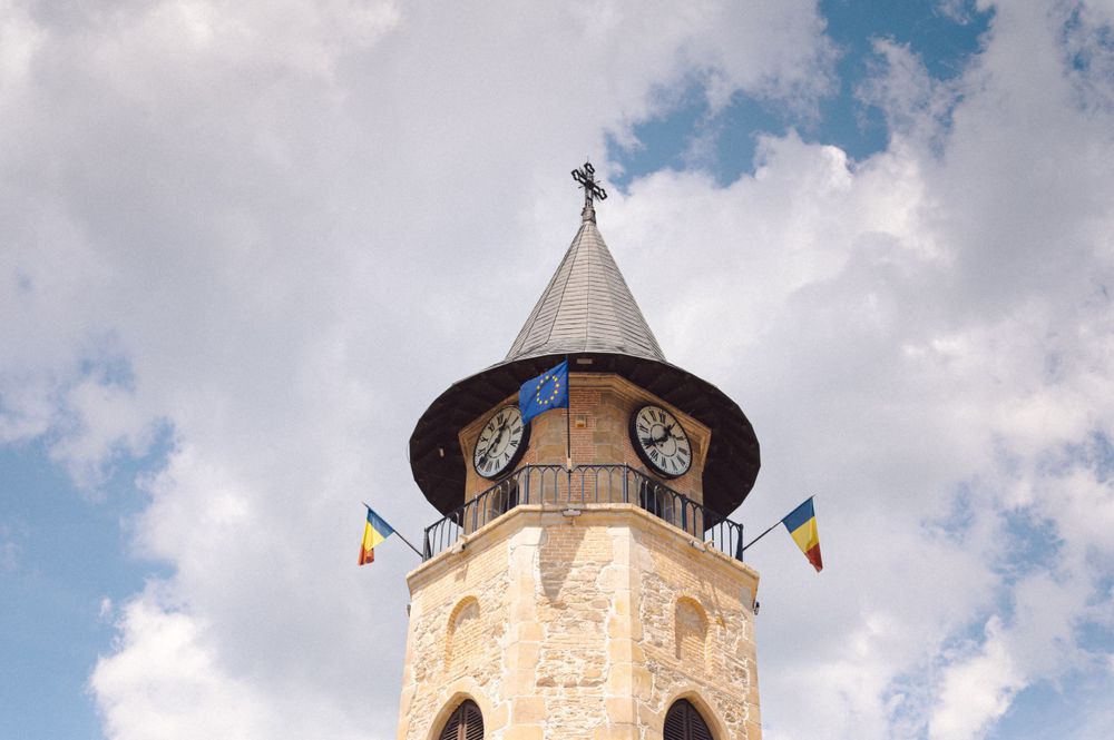 Stone church tower with clock and pointed spire against blue sky with white clouds.