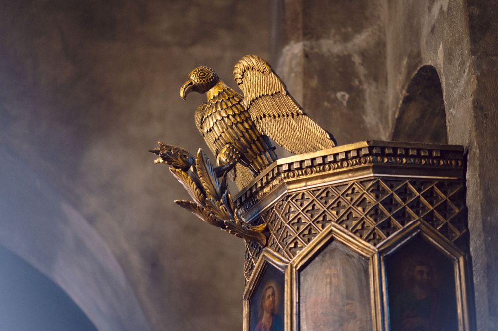 A golden eagle sculpture perches on an ornate Gothic architectural detail in dramatic lighting.