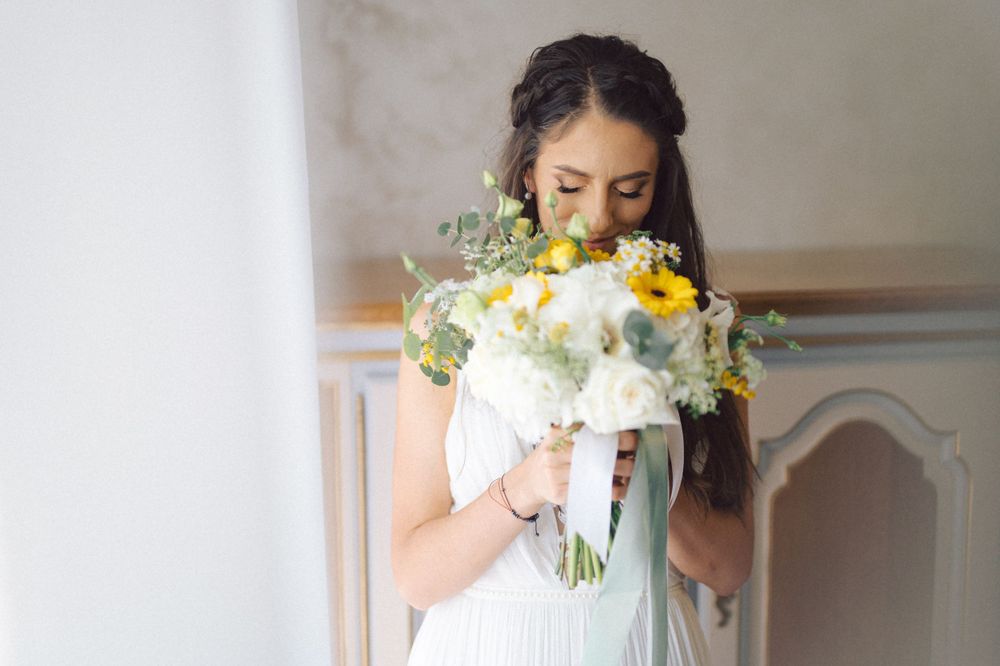 Person in white wedding dress smelling a bouquet of sunflowers and white flowers.