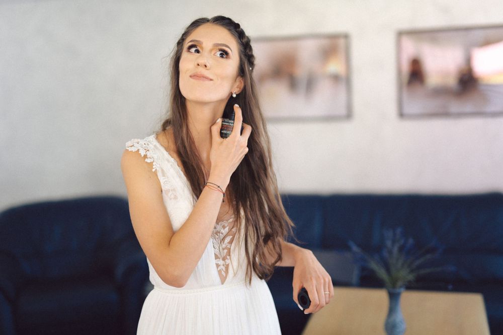 A woman in a white lace dress applies perfume with an upward glance in a room with framed artwork.