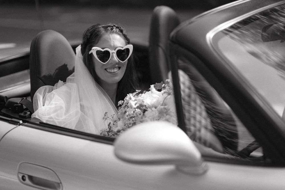 A convertible car carries a bride wearing stylish sunglasses in black and white photography.