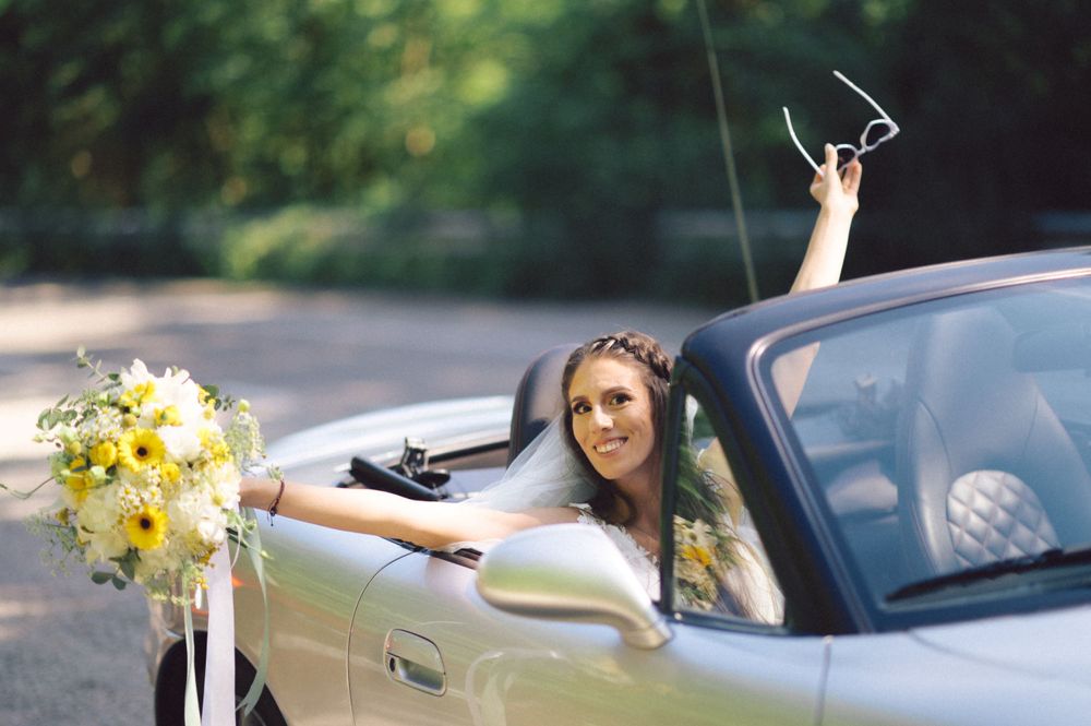 A bride in a convertible car holds up a yellow bouquet while celebrating.