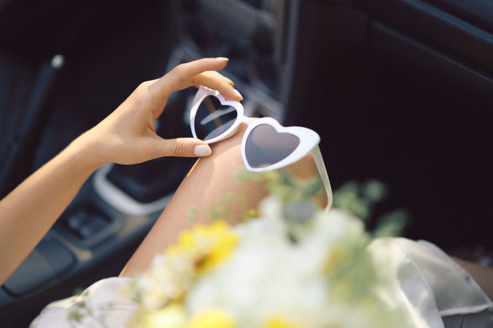 Hand holding heart-shaped sunglasses against a blurred car interior background.
