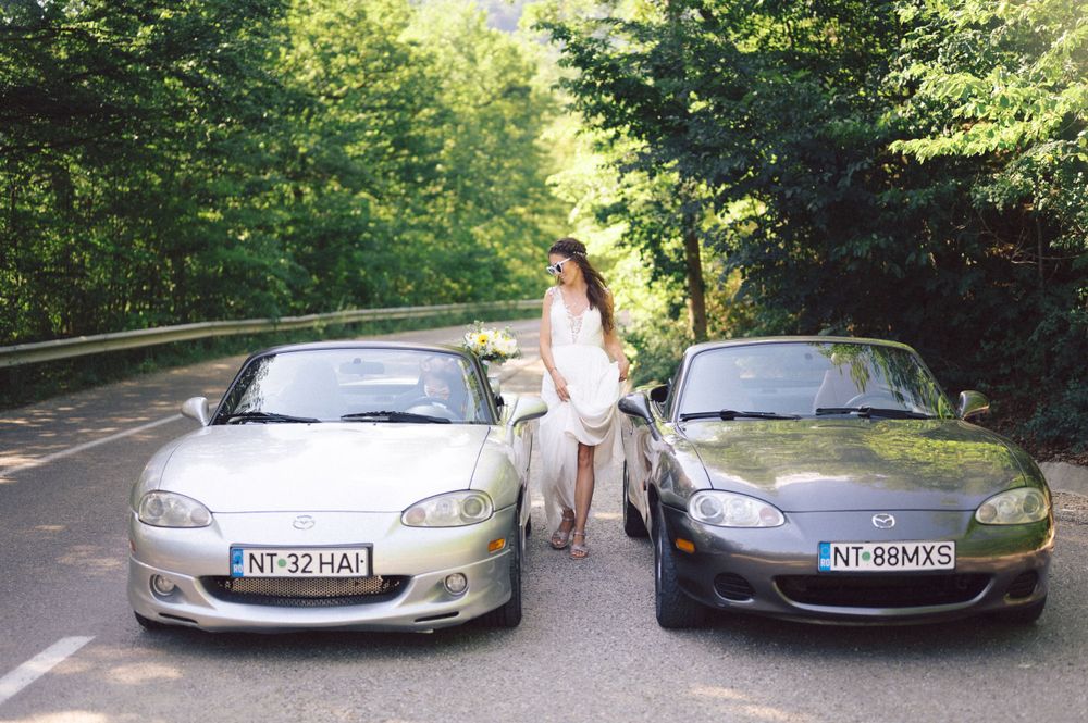 Two silver sports cars parked on a tree-lined road with a bride in white between them.