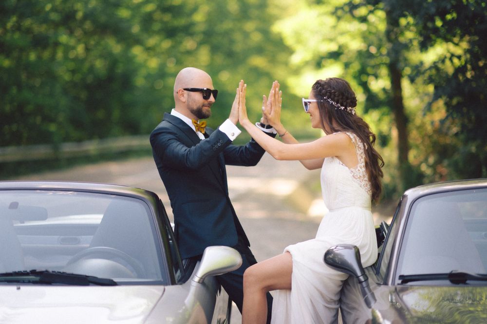 A playful wedding photoshoot with a couple high-fiving between two parked cars on a tree-lined road.