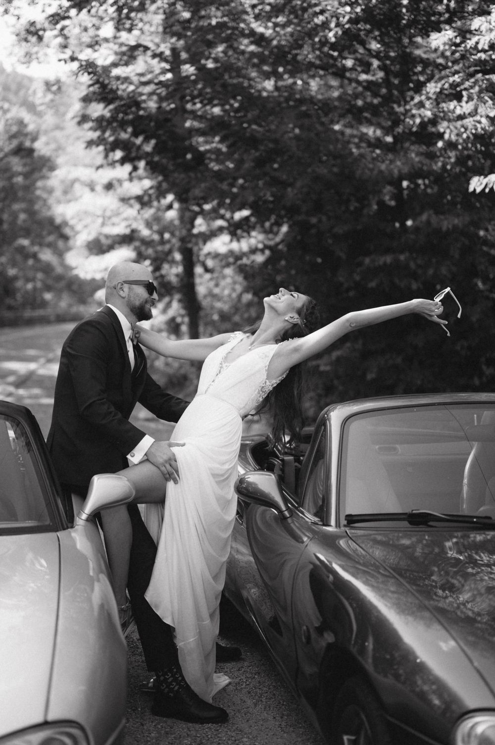 An artistic black and white wedding photo showing a dramatic pose between two vintage cars.