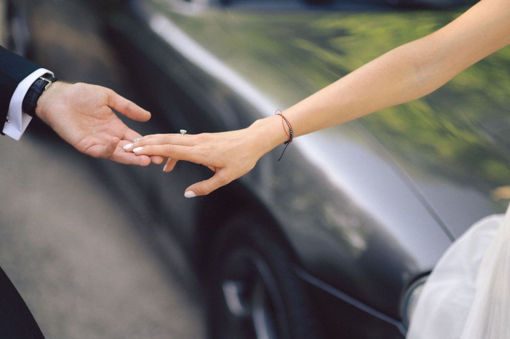 Close-up shot of hands reaching toward each other near a car during a wedding photoshoot.