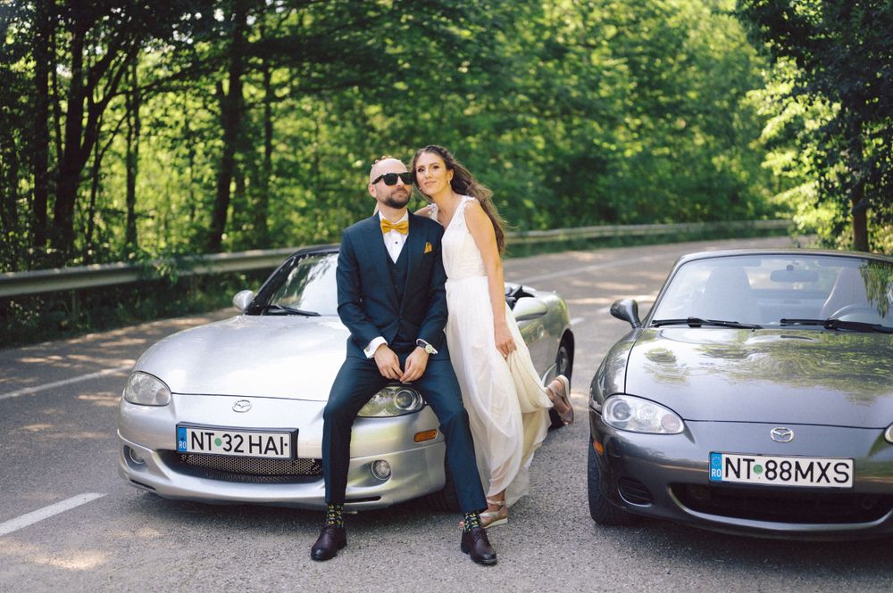 Wedding couple posing together between two silver sports cars on a forest road.