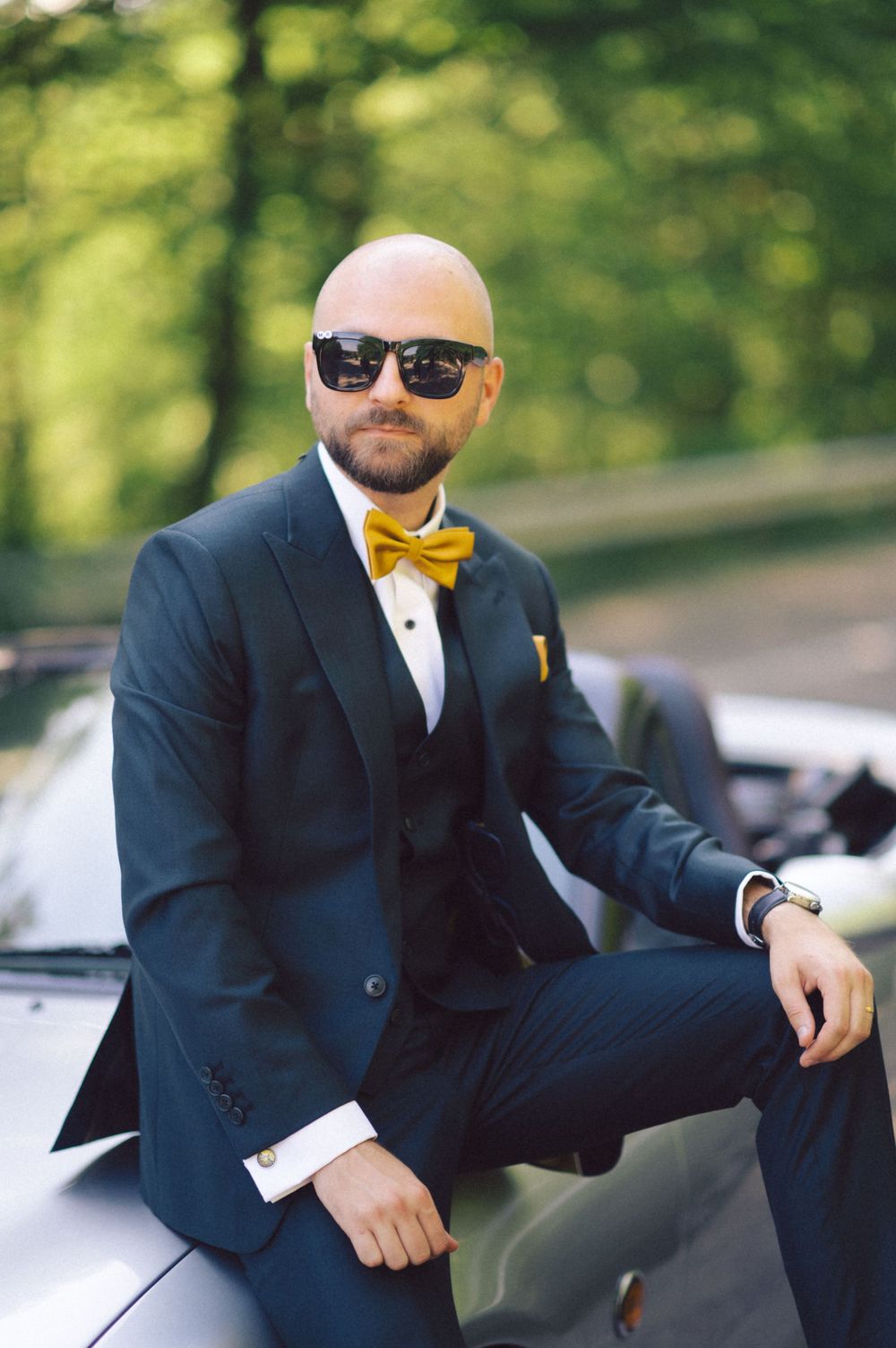 Stylish groom in navy suit and yellow bowtie poses on silver car during wedding photoshoot.