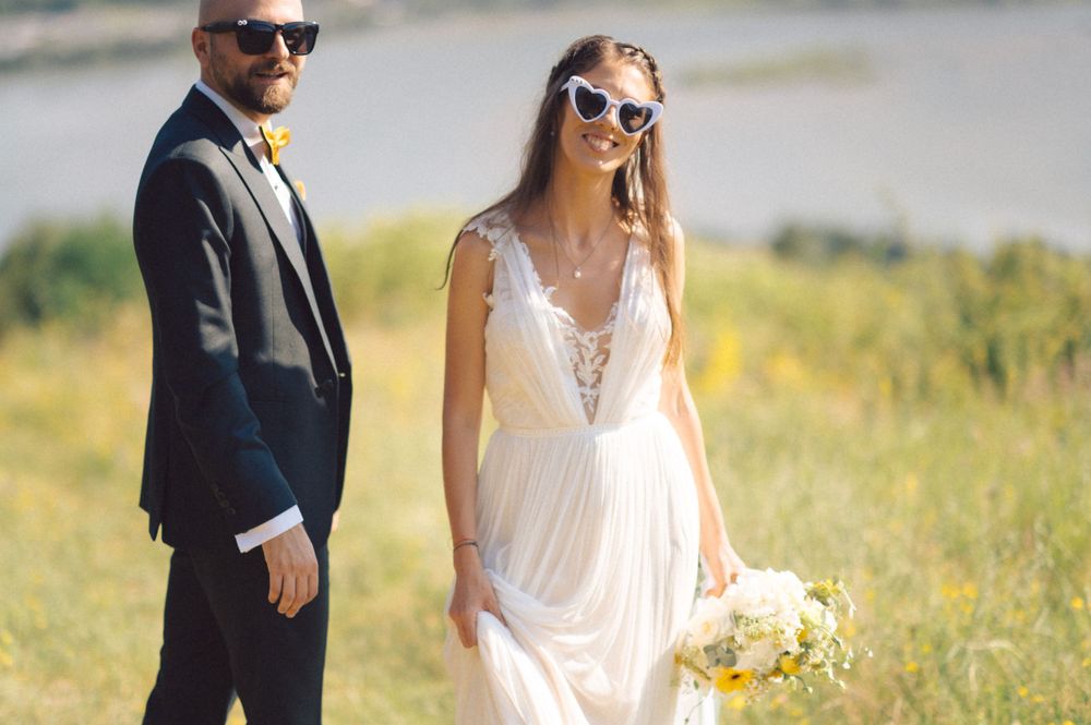 Newlyweds in sunglasses pose in a sunny field with a white bouquet against a natural backdrop.