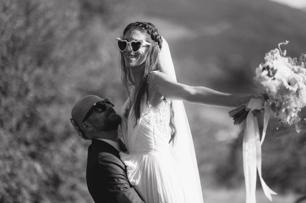 Black and white photo of playful wedding couple posing with sunglasses outdoors.