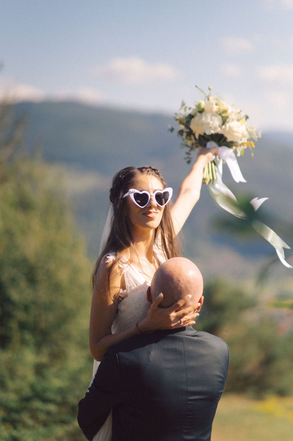 Bride in heart-shaped sunglasses holds bouquet while celebrating on mountainside.