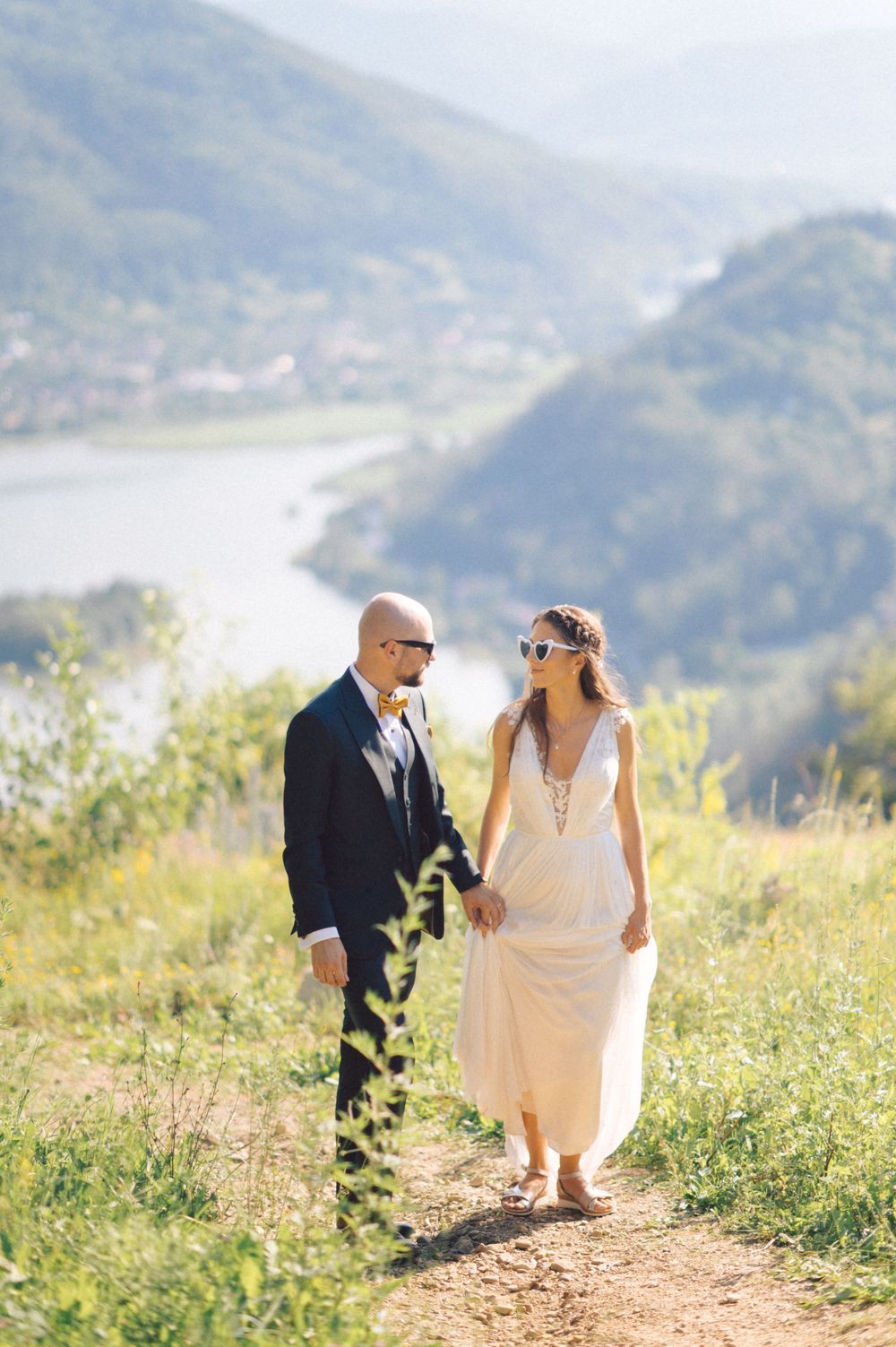 A wedding couple walking hand in hand on a hilltop trail overlooking a scenic river valley.
