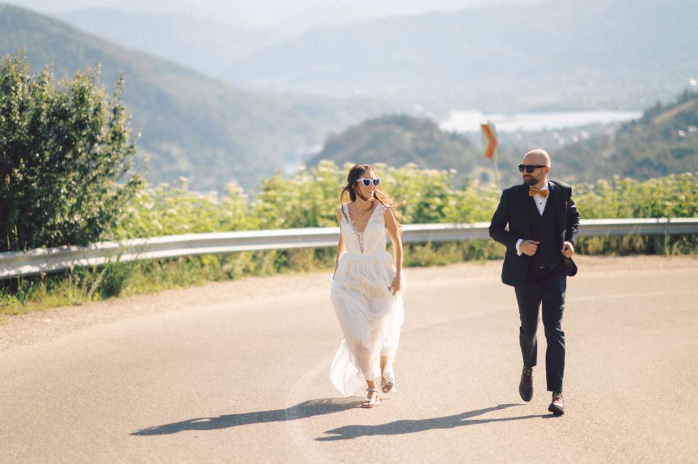 A wedding couple running playfully on a mountain road with scenic valley views.