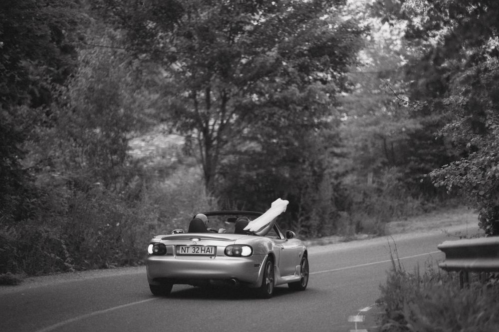Black and white photo of a vintage convertible car driving on a winding forest road.