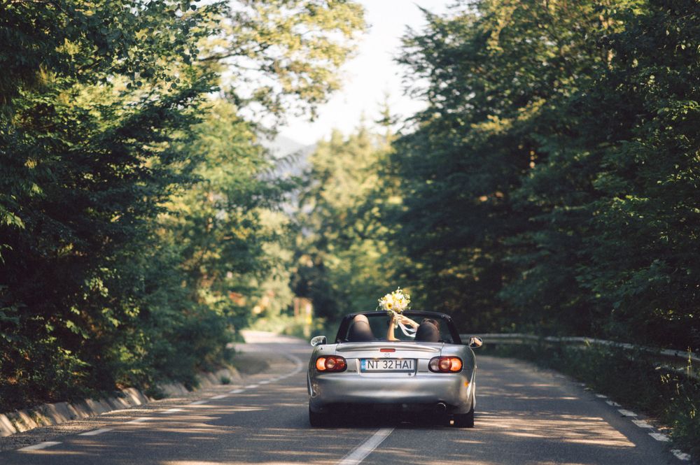 A silver convertible car driving down a tree-lined mountain road on a sunny day.