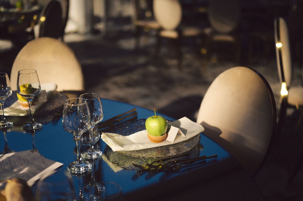 Formal place setting with blue tablecloth featuring wine glasses and a decorative green apple centerpiece.
