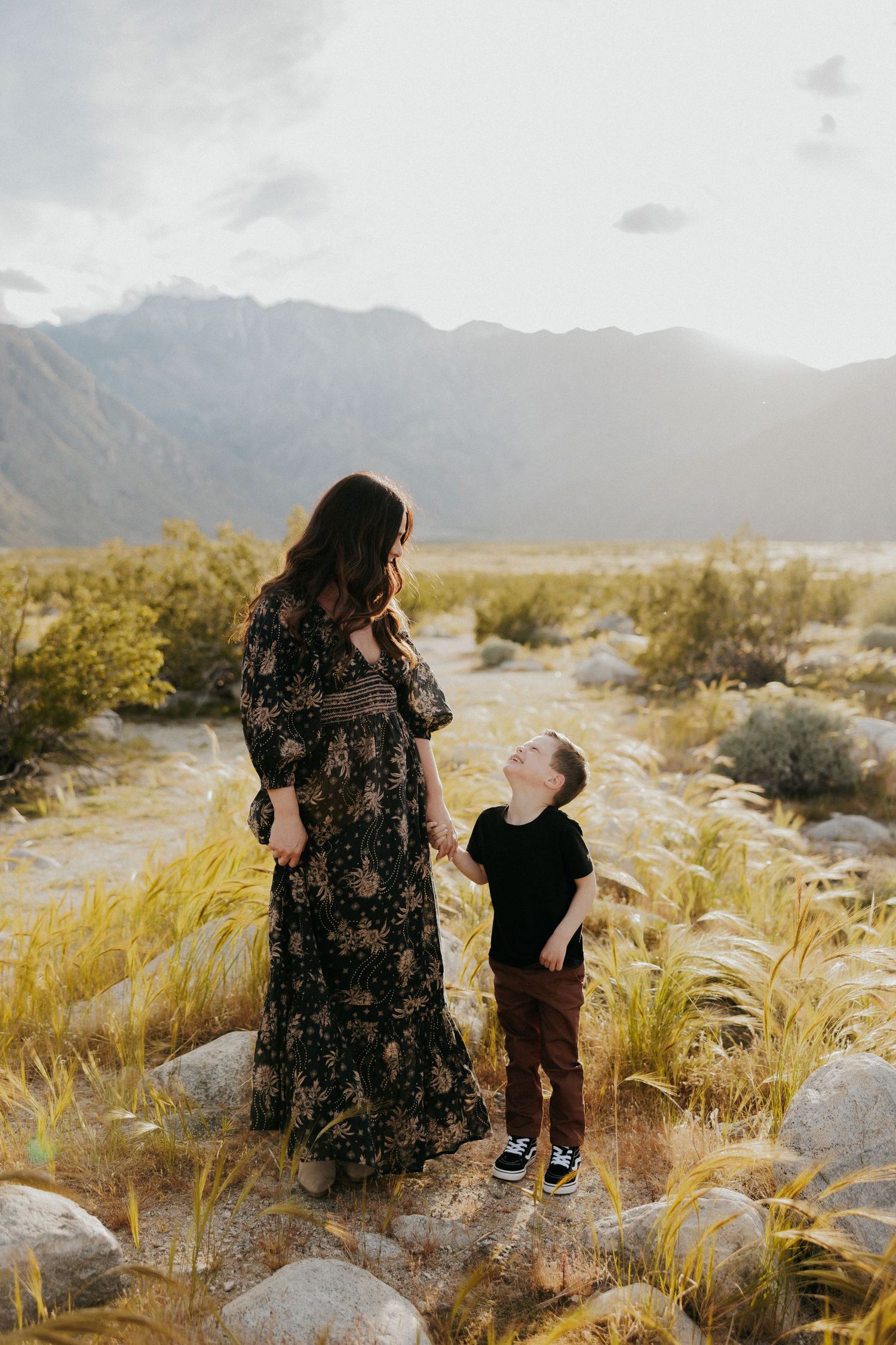 A woman in a floral maxi dress and child in black shirt stand together against mountain backdrop during golden hour.