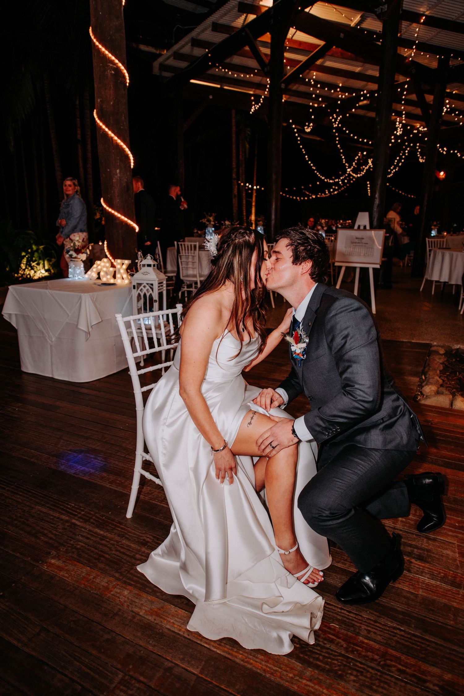 Couple performs traditional garter removal during wedding reception on rustic wooden dance floor.