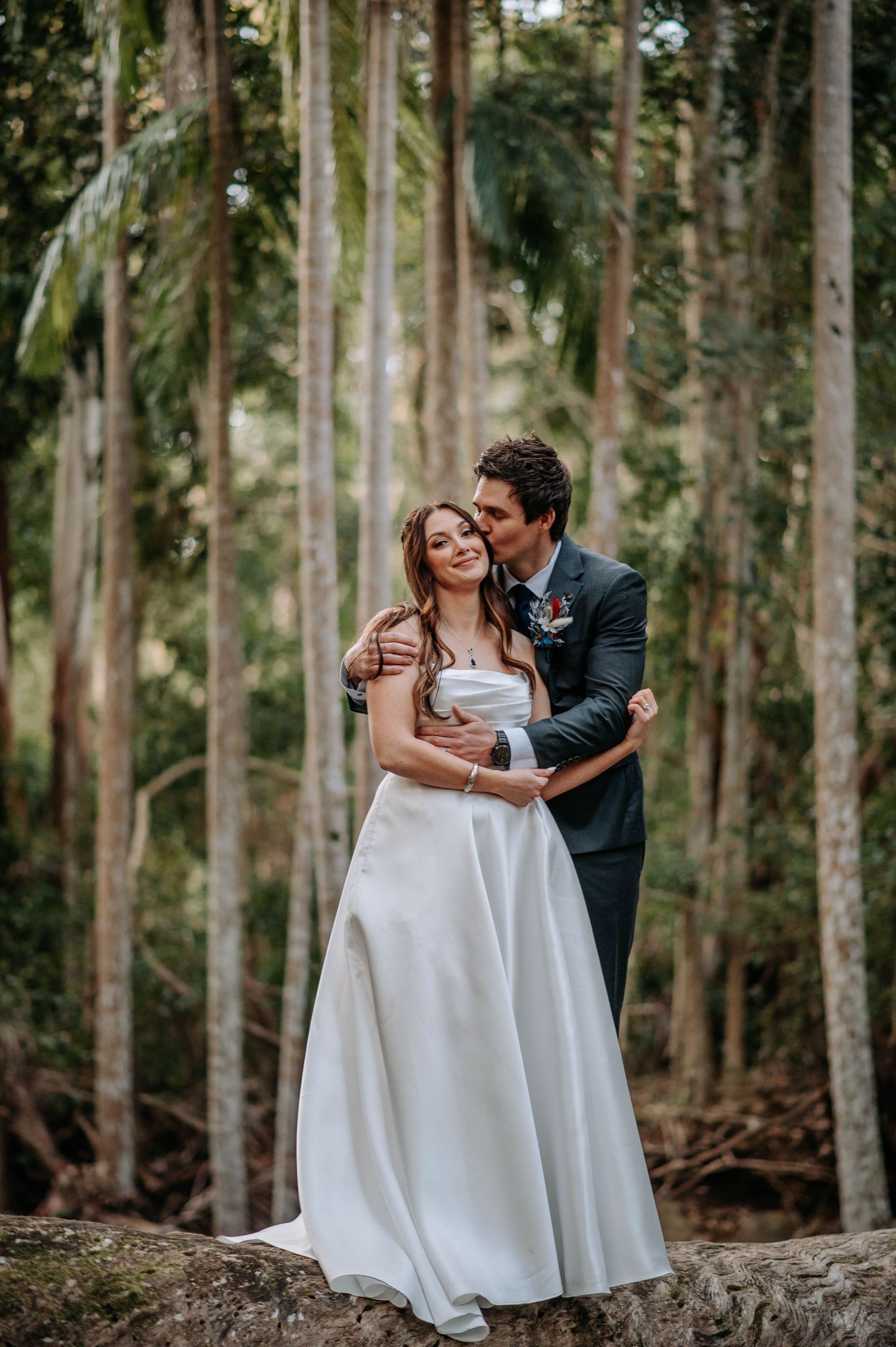 A romantic wedding portrait of a couple embracing on a log in a lush forest setting.