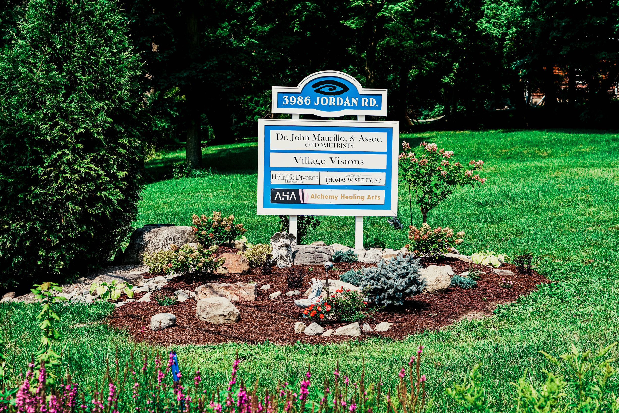 Blue business sign with white lettering surrounded by landscaped garden bed and flowers.