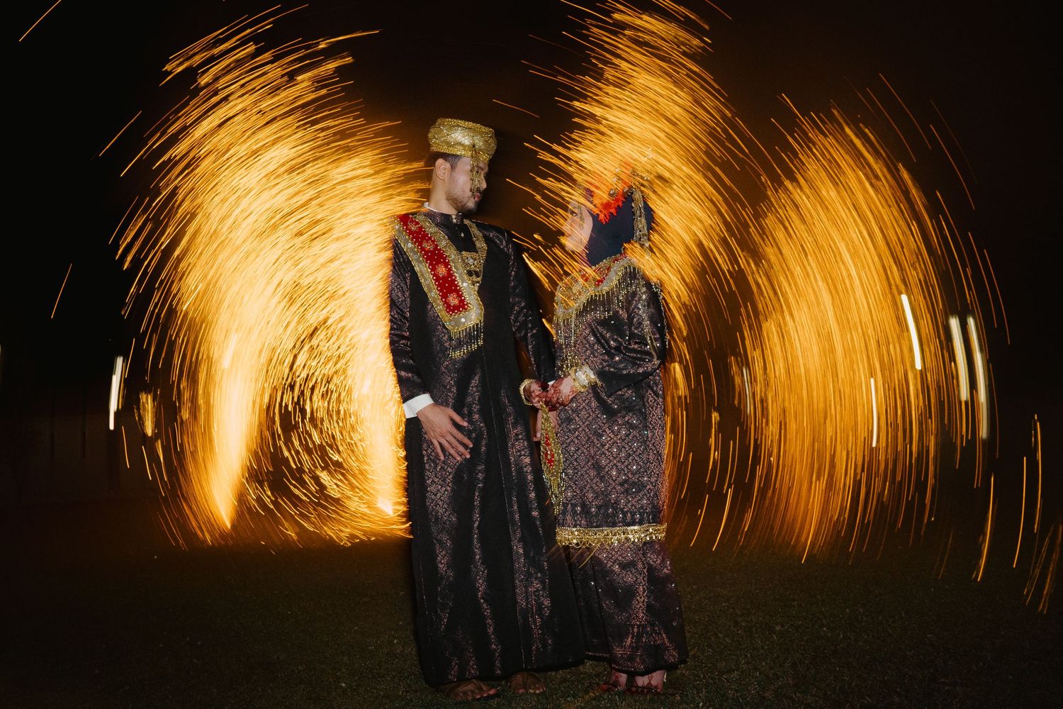 Couple in traditional Indonesian wedding attire posing with golden fireworks spinning around them at night.