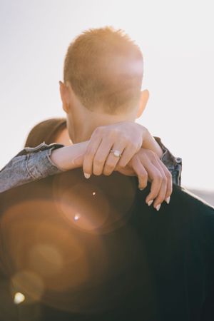 A person wearing a wedding ring embraces another in silhouette against bright sunlight with lens flare effects.