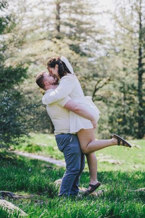 A romantic couple embraces joyfully in a sunlit forest setting during what appears to be an engagement photo session.