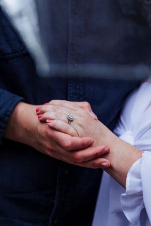 Two people holding hands tenderly, with one wearing a dark jacket and the other in white, showing a silver ring.