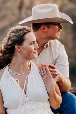 A romantic couple in white outfits pose outdoors, with one wearing a cowboy hat and holding hands in warm sunlight.