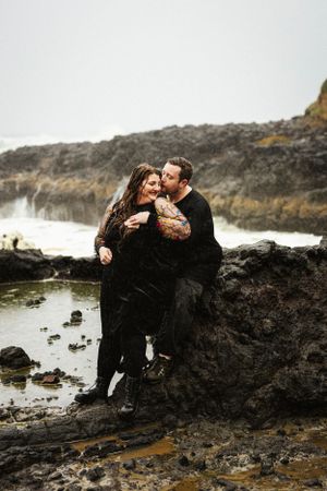 Couple embraces on rocky coastline with crashing waves in background.