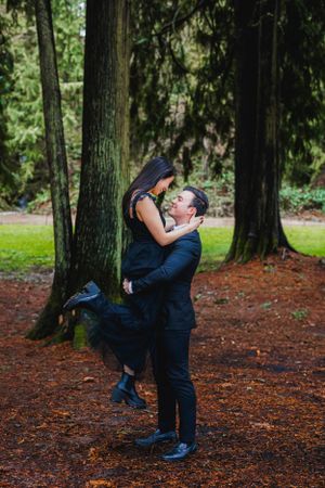 A romantic couple embraces in a forest setting with tall trees and fallen autumn leaves on the ground.