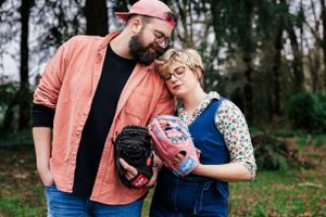 A couple stands laughing outdoors in casual attire while holding baseball mitts and a colorful ball.
