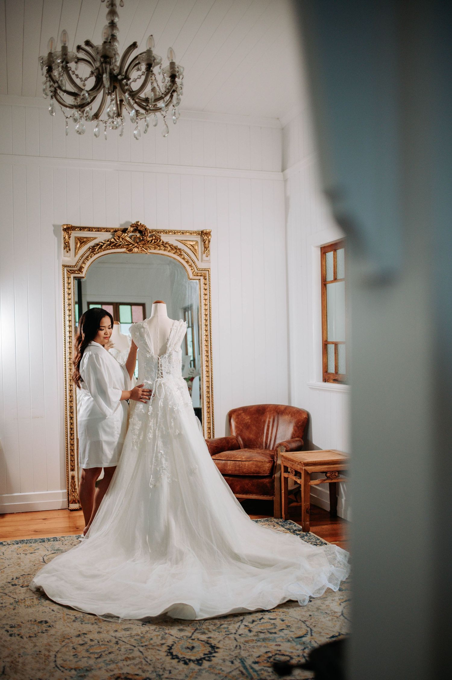 An elegant white wedding dress displayed in a room with an ornate gold mirror and chandelier.