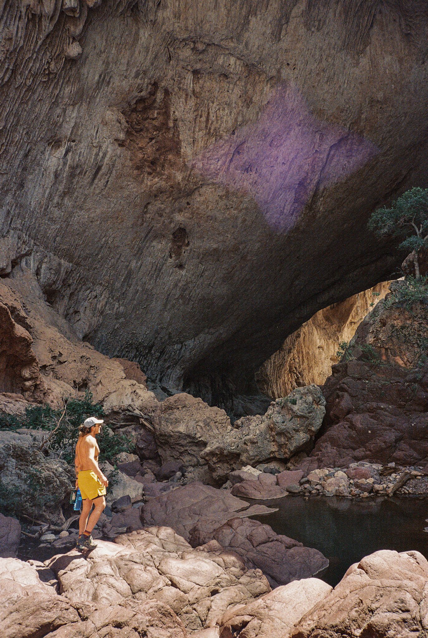 Explorer in orange vest stands at edge of rocky cave formation with purple-lit ceiling.