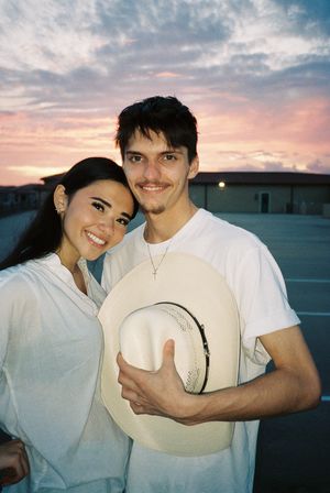 Playful couple posing with a cowboy hat against a sunset sky.