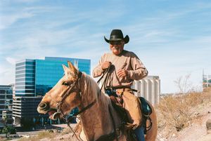 Horseback rider in cowboy hat against modern office building backdrop.