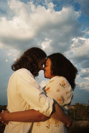 Two people embrace against a cloudy sky backdrop wearing light colored clothing.