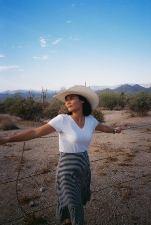 Person in western attire posing playfully against a desert landscape with mountains and cacti.