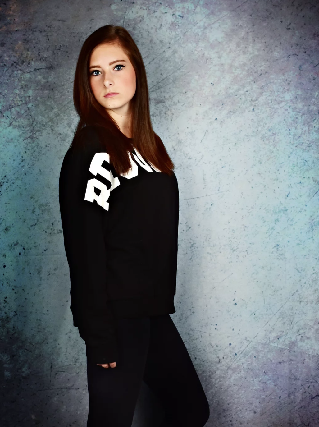 Young woman with red hair poses in black sweatshirt with white lettering against blue-gray studio background.