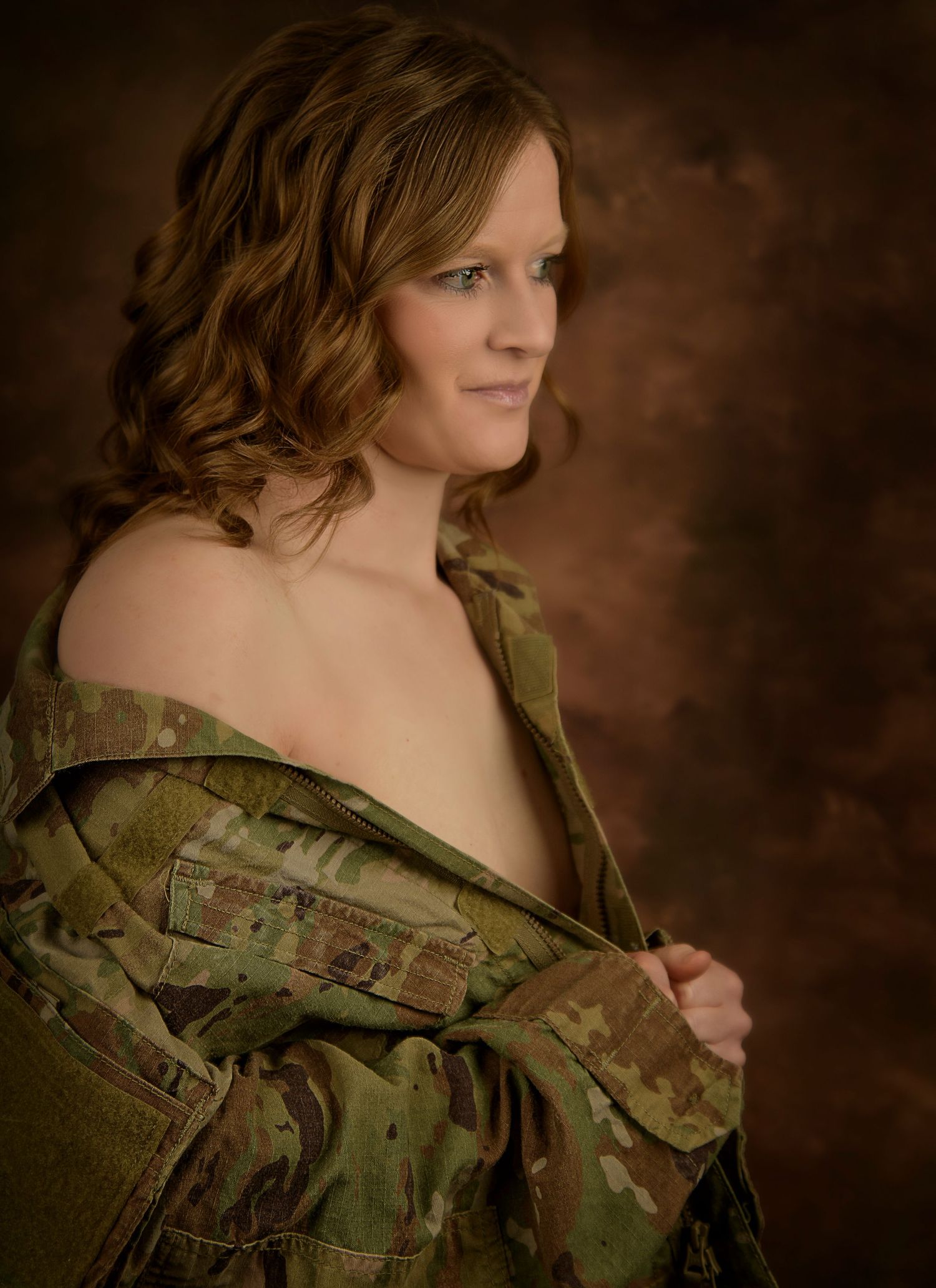 Woman in camouflage military jacket posing against brown studio backdrop with curly auburn hair.