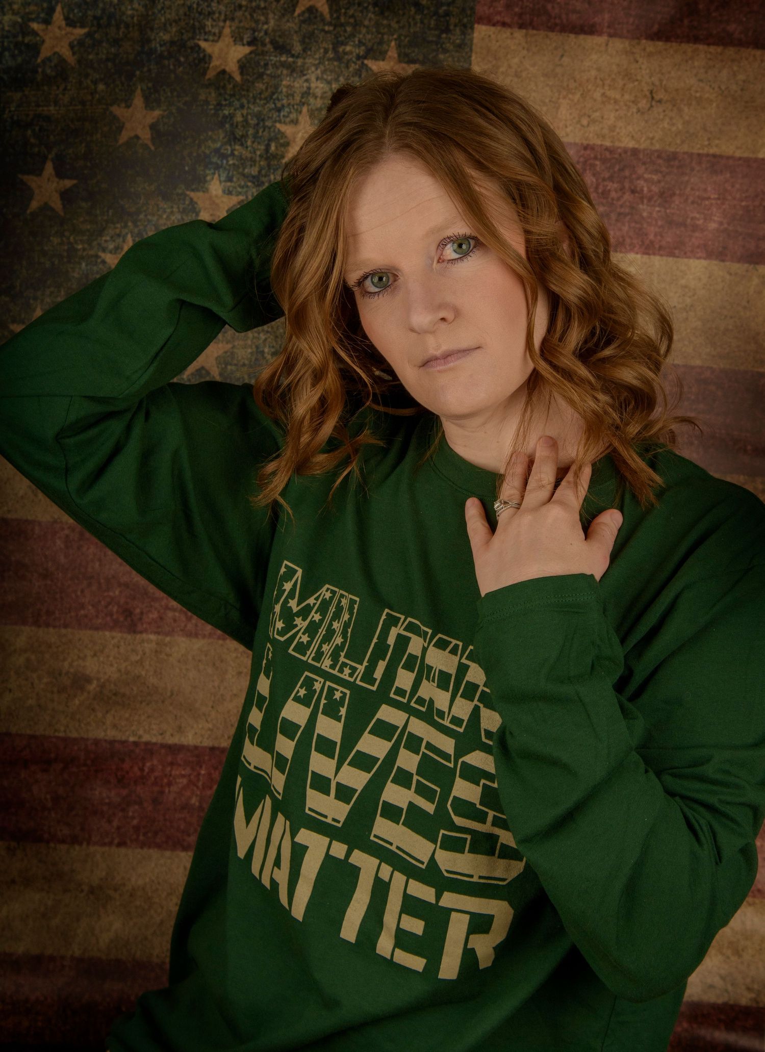 Young woman with curly red hair wearing a green Military Lives Matter sweatshirt posed against an American flag background.