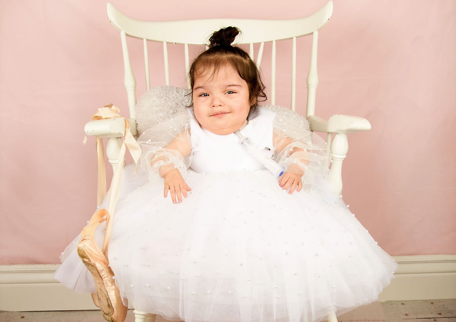 A cute baby girl in a white tutu dress sits smiling in a white chair against a pink background.
