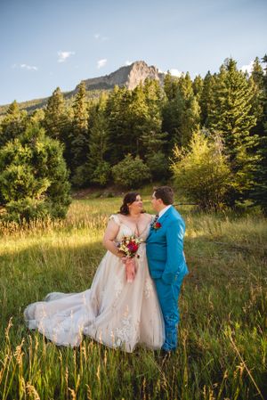 Couple shares romantic moment in field with dramatic mountain peak and forest scenery.