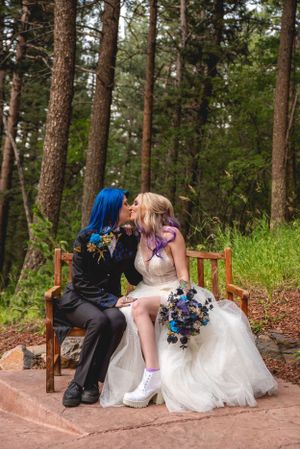 Newlyweds share an intimate moment on a wooden bench surrounded by pine trees.