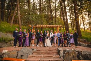 Wedding ceremony group photo under string lights and wooden arch in pine forest.